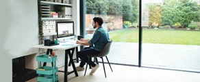 Man sat on chair at desk looking at his phone next to a window with a printer, folders and plants on shelving
